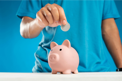 A person is inserting a coin into a pink piggy bank, symbolizing savings and financial planning for situations like postal disability.