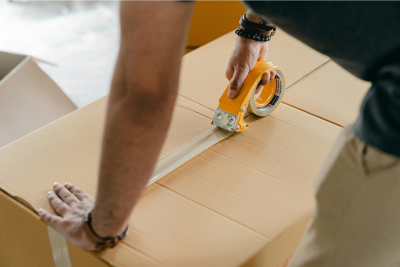 Close-up of a person’s hands using a yellow tape dispenser to seal a cardboard box, representing postal paycheck protection.